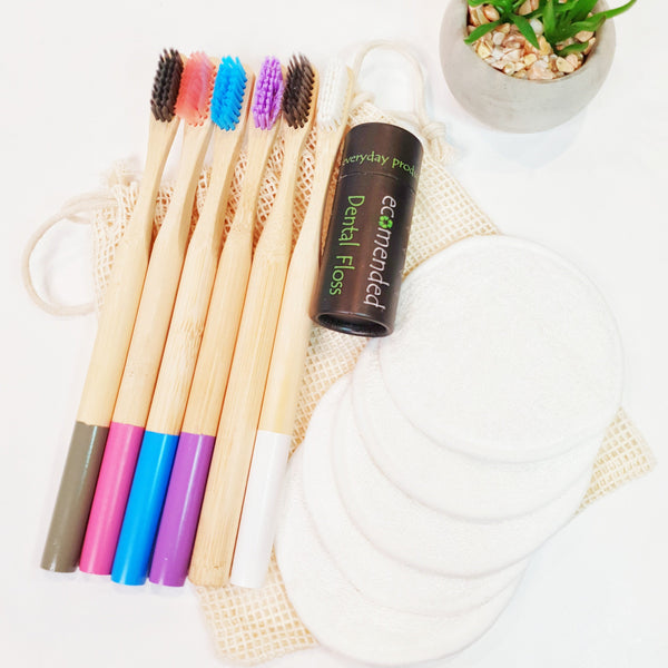 6 different coloured bamboo toothbrushes with bamboo dental floss and 5 white reusable makeup remover pads on mesh laundry bag.